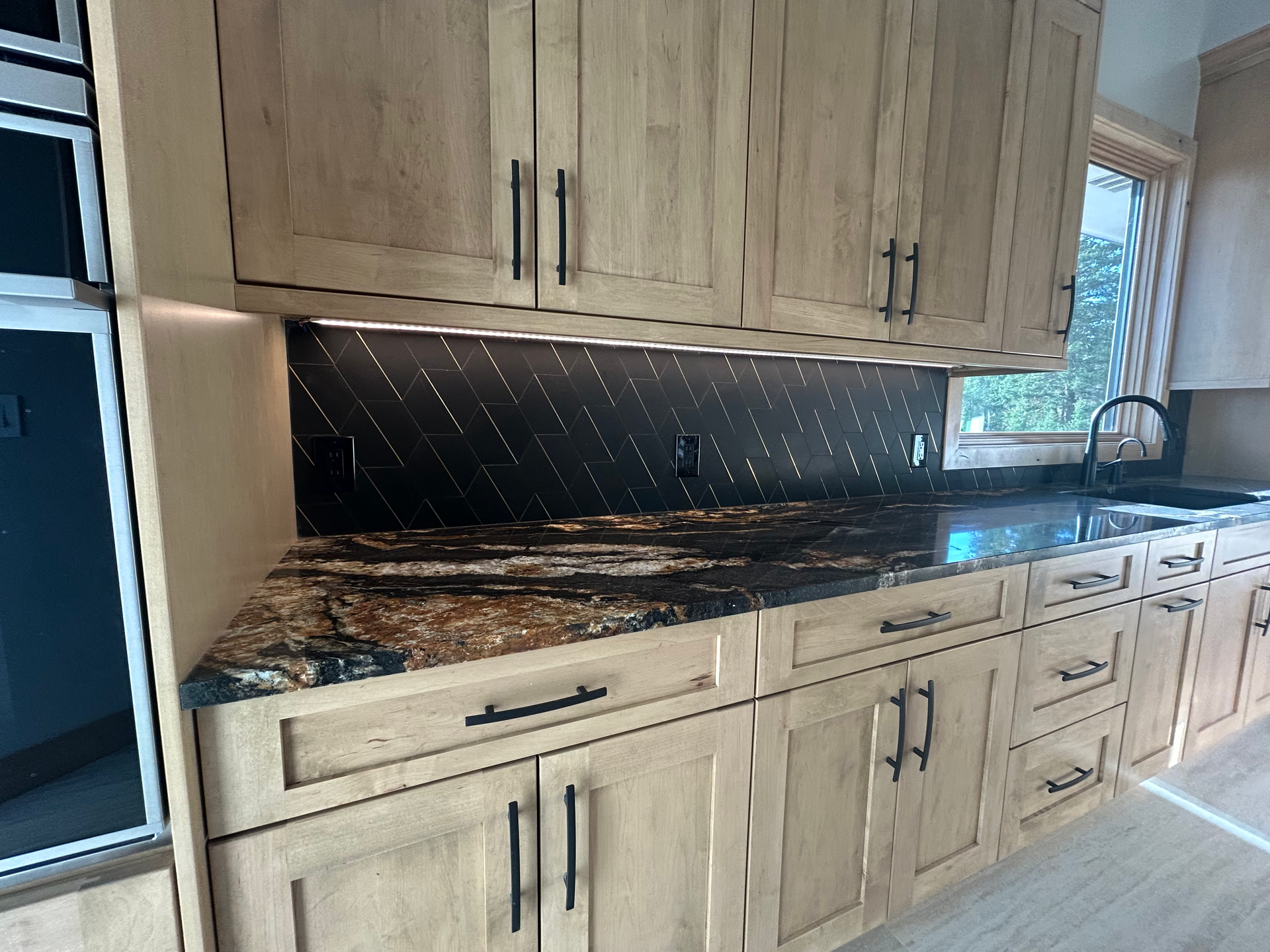 Kitchen with light wood cabinets, black countertops, dark backsplash, and black hardware.
