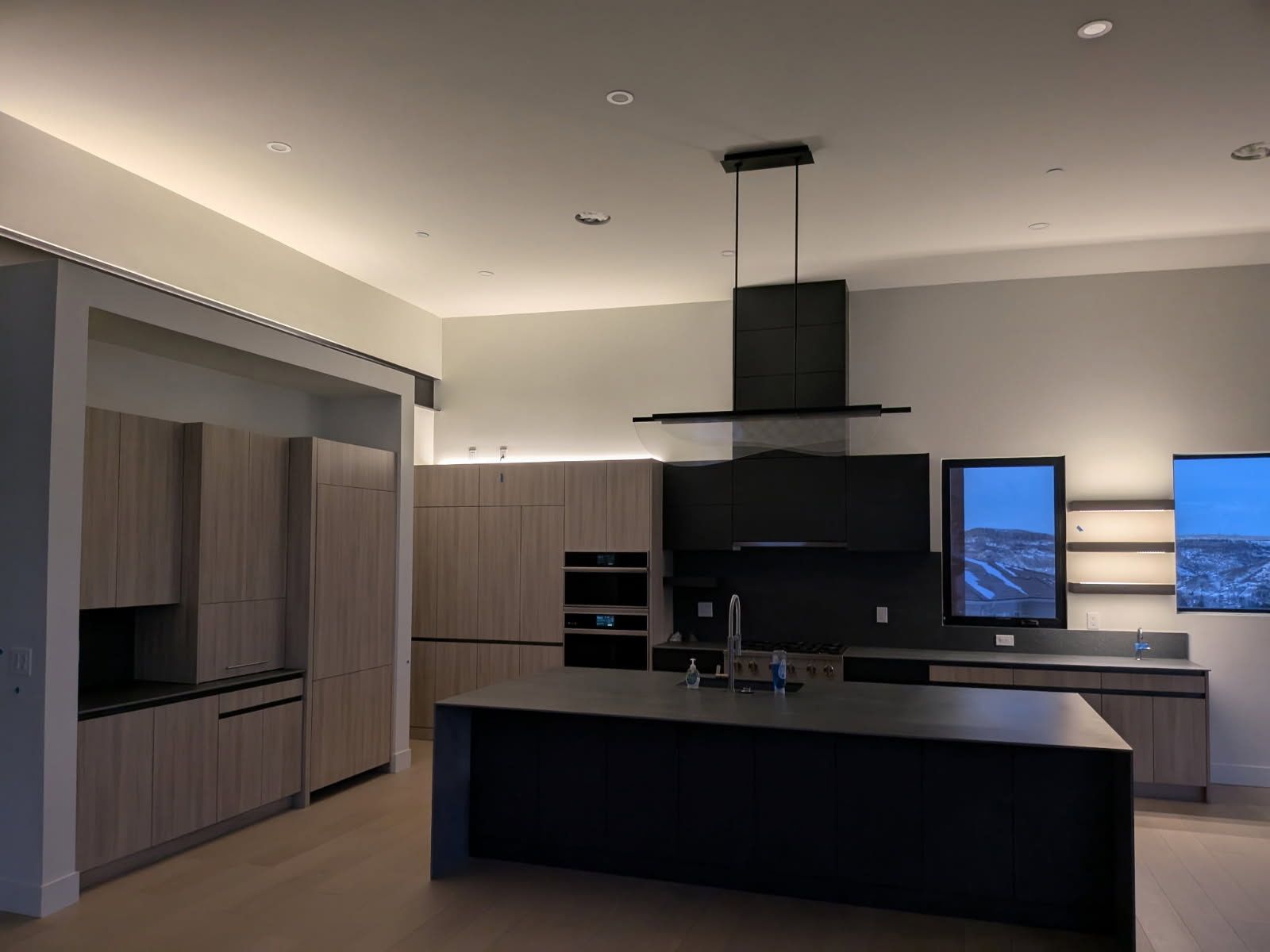Modern kitchen with light wood cabinets, black island, and range hood; lit by recessed and strip lighting.