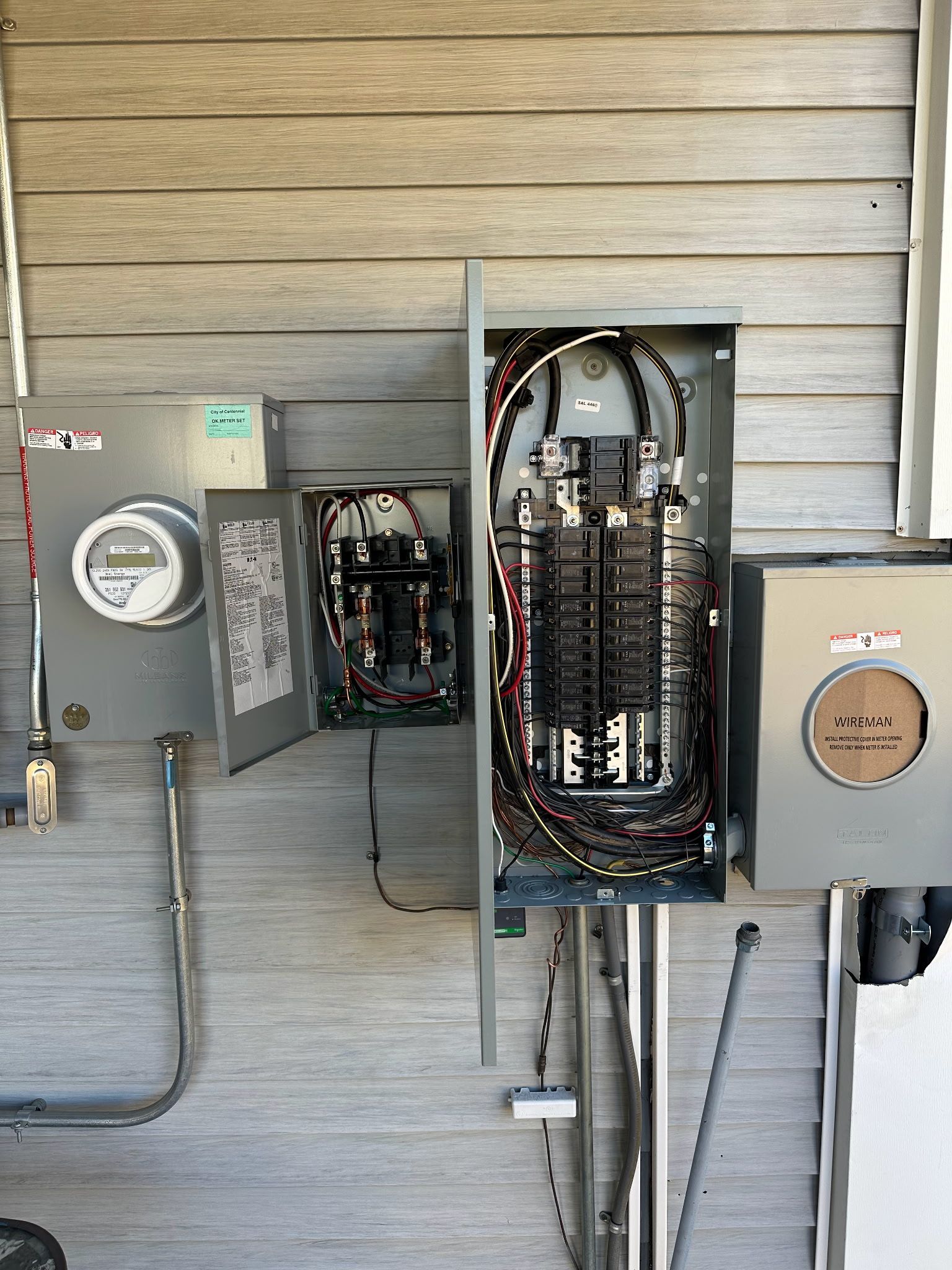 Electrical panel boxes mounted on a gray exterior wall, with open panel showing wiring.