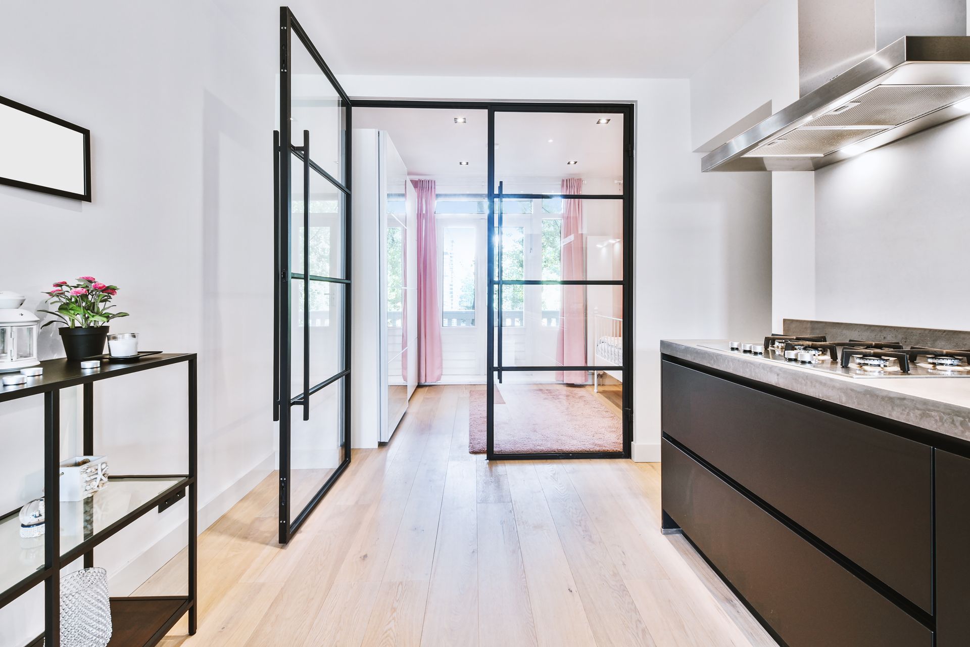 Modern kitchen with black framed glass doors leading to a room with pink curtains; black cabinet and steel range hood.