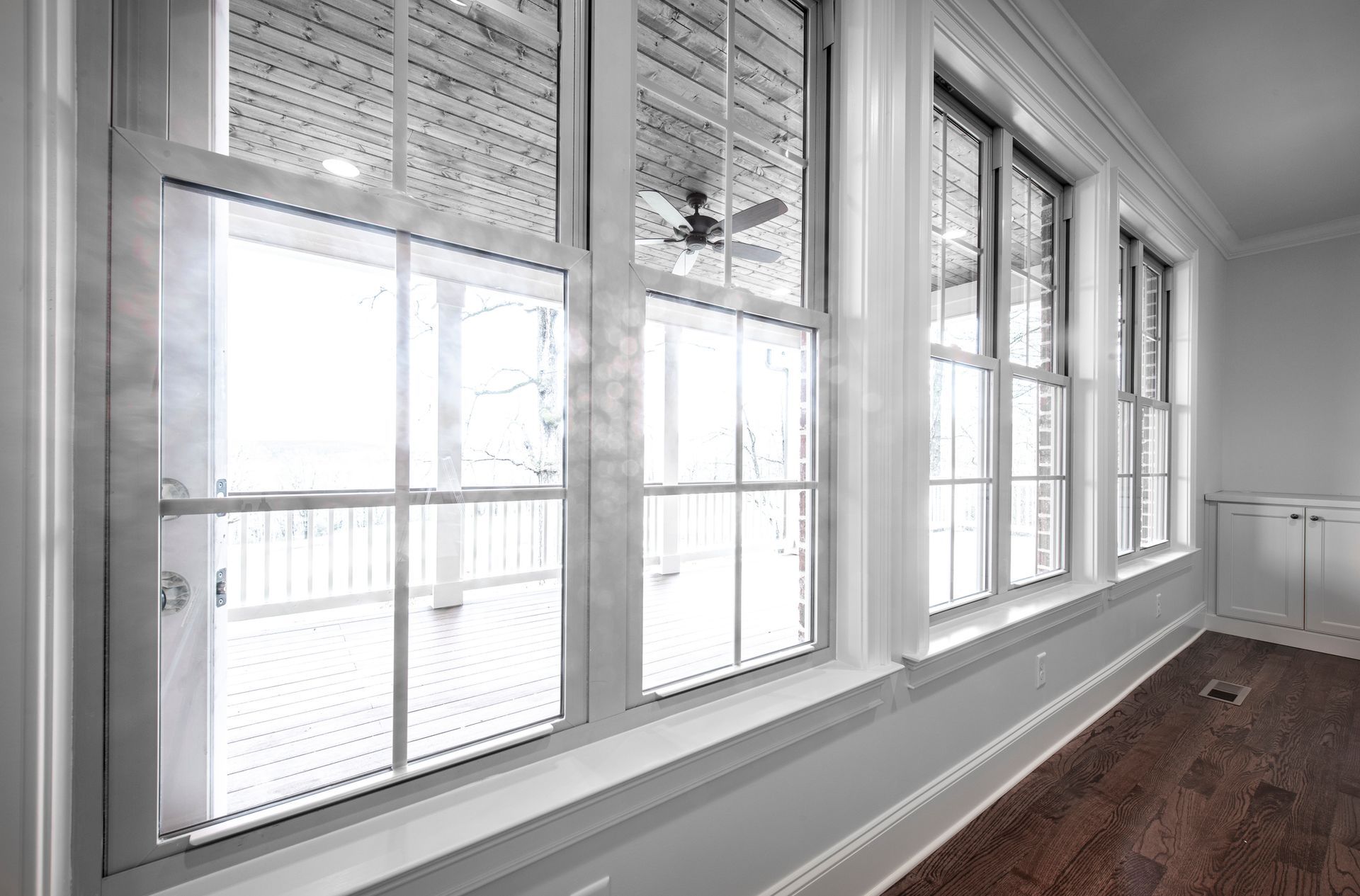 Row of tall, white-framed windows overlooking a balcony with a ceiling fan; interior with wood floor, white walls.