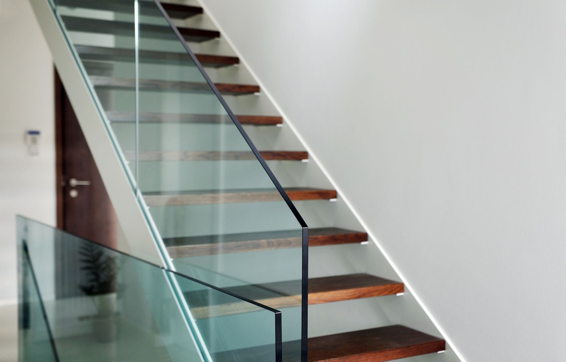 Wooden staircase with glass railing and black handrail against a white wall.