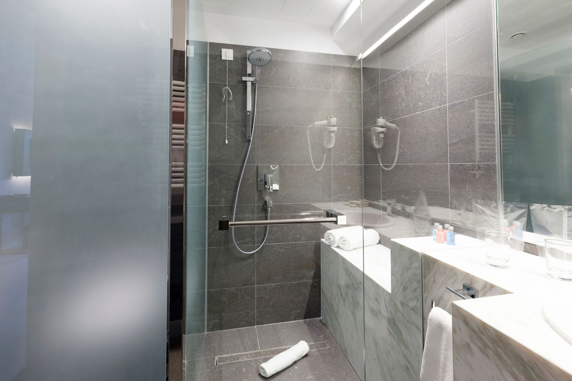 Shower stall with gray tile and glass door, towel on the floor, and sink in a modern bathroom.
