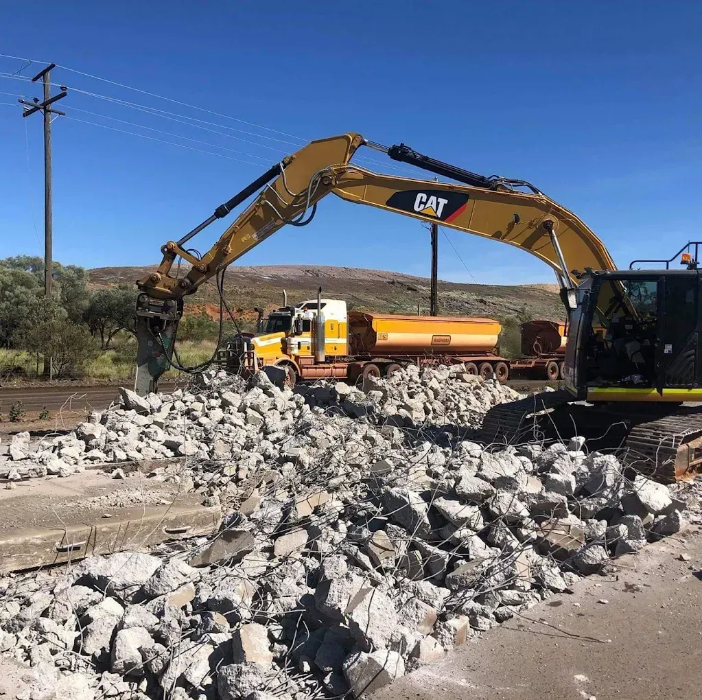 Yellow excavator demolishing concrete road, with a truck in the background — Lamont Civil Services Pty Ltd In Townview, QLD