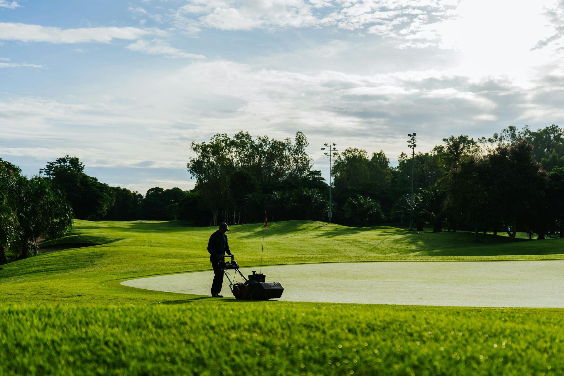 A silhouette of a person uses a push mower to maintain a vibrant green golf course under a cloudy sky.