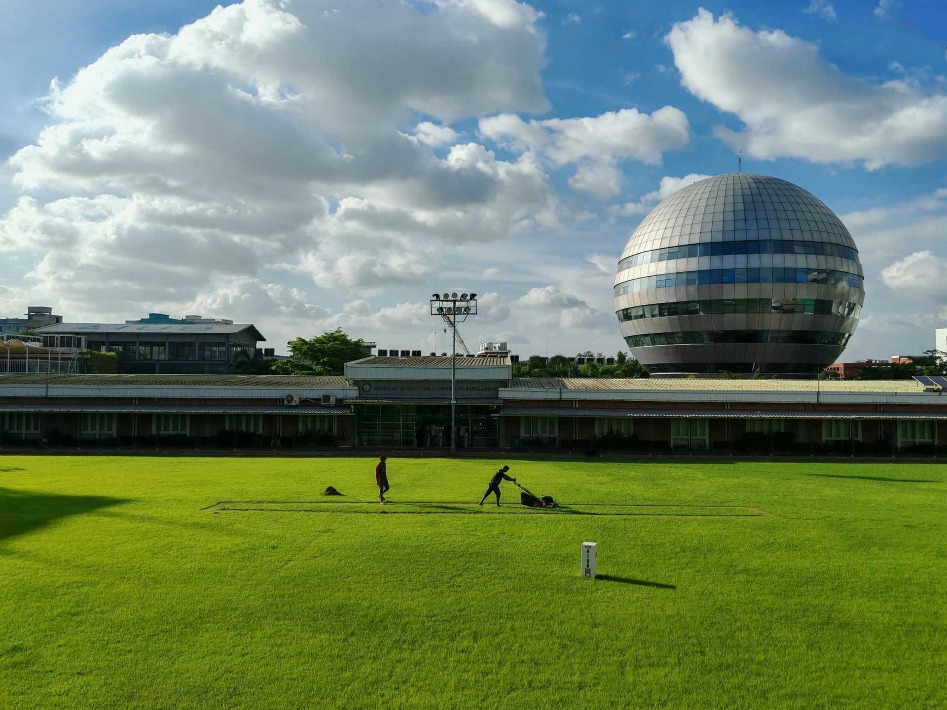 Two people tend to a bright green lawn before a large, silver sphere building under a blue, cloudy sky.