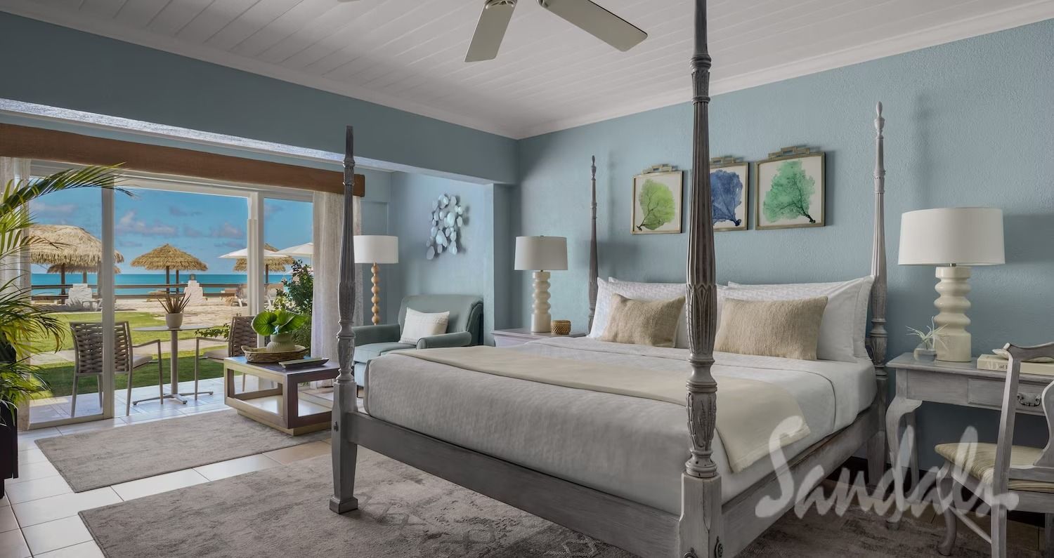 Hotel bedroom with a view of the beach through sliding glass doors. Light blue walls, a four-poster bed, and white furniture.