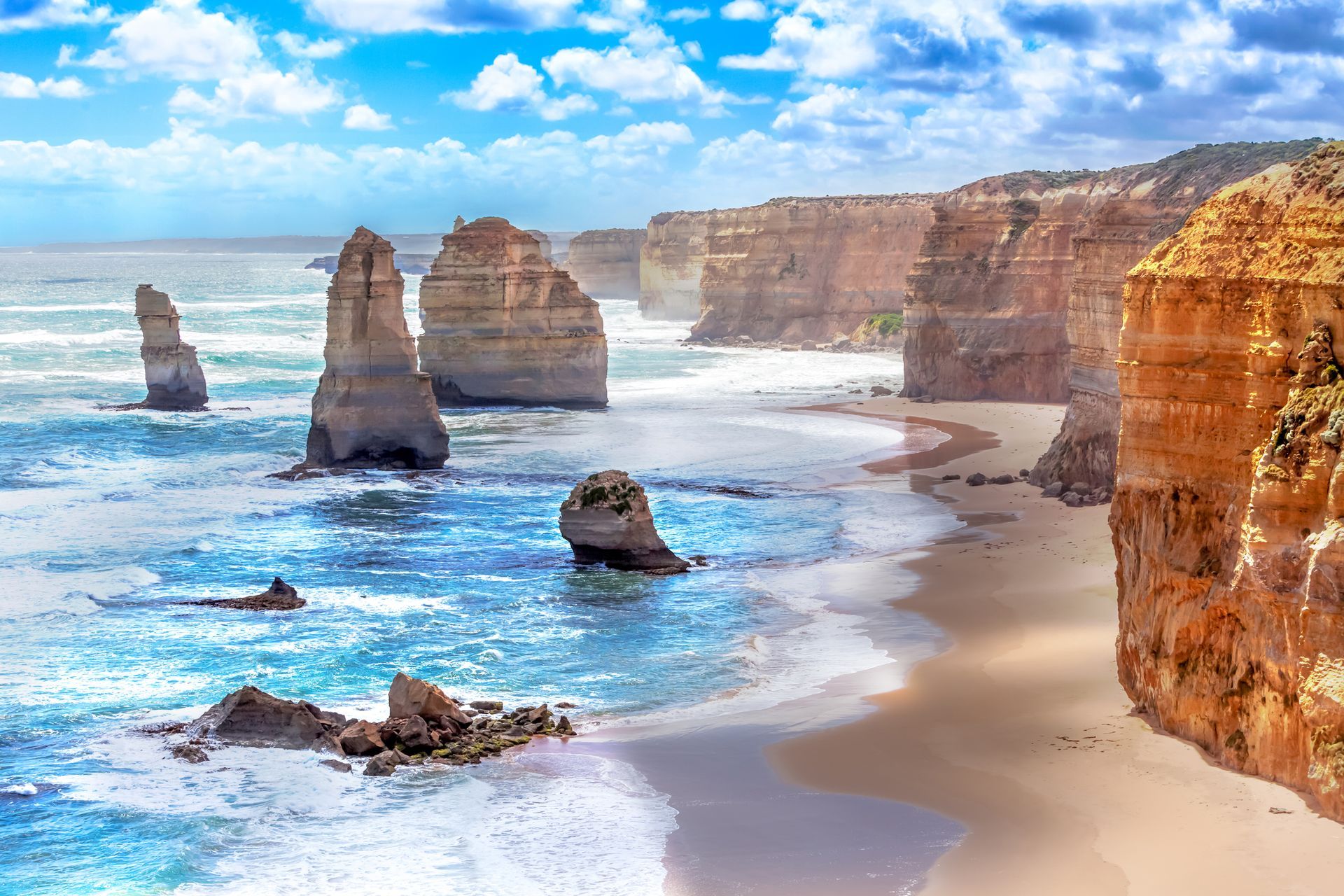 Coastal cliffs and sea stacks along a beach with turquoise water under a blue sky.