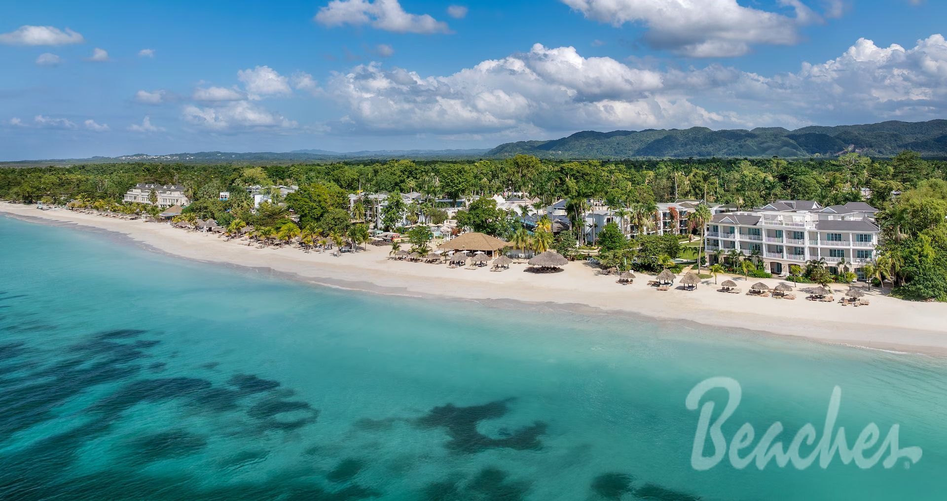 Aerial view of a white sandy beach, turquoise water, and a resort with palm trees.