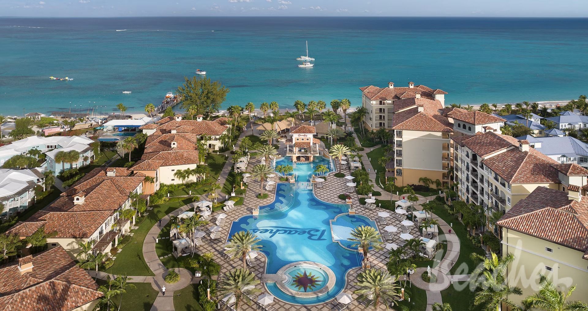 Aerial view of a resort with a large pool and buildings, next to a turquoise ocean.