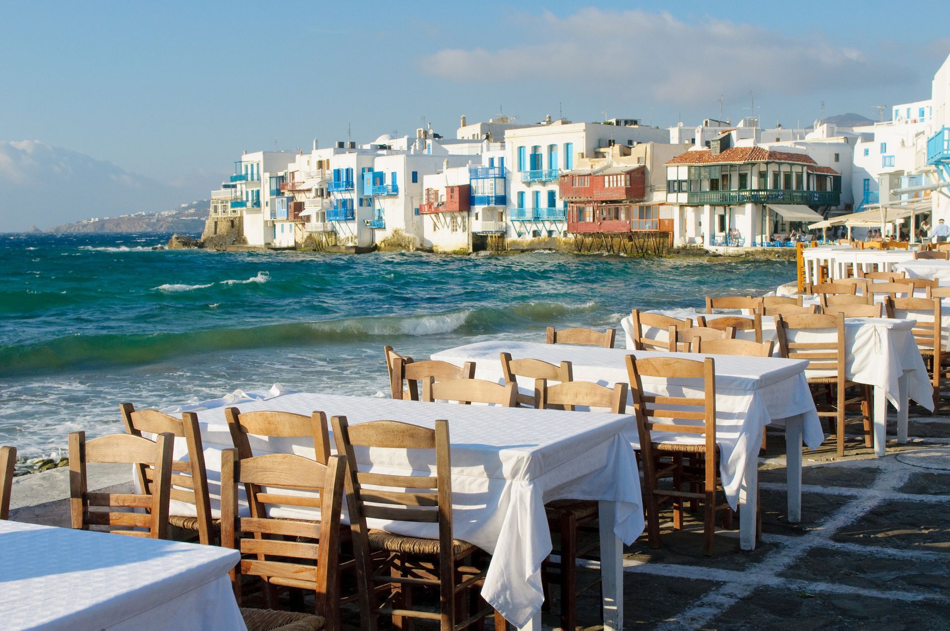 Restaurant tables set on a cobblestone patio overlooking the sea and colorful buildings in Mykonos, Greece.