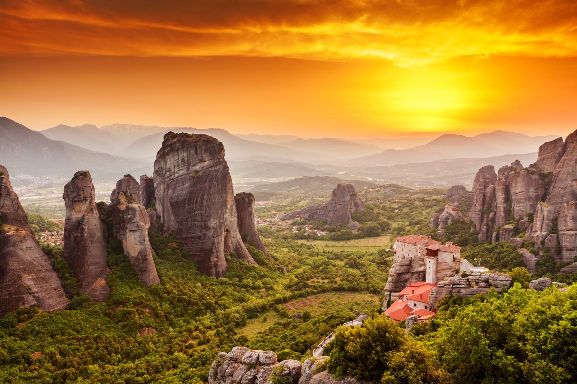 Sunset over Meteora monasteries in Greece, with rocky formations and vibrant orange sky.