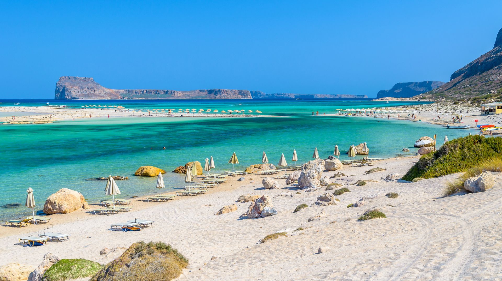 Turquoise water beach, white sand, light-colored umbrellas, large rock formations. Distant island under clear blue sky.