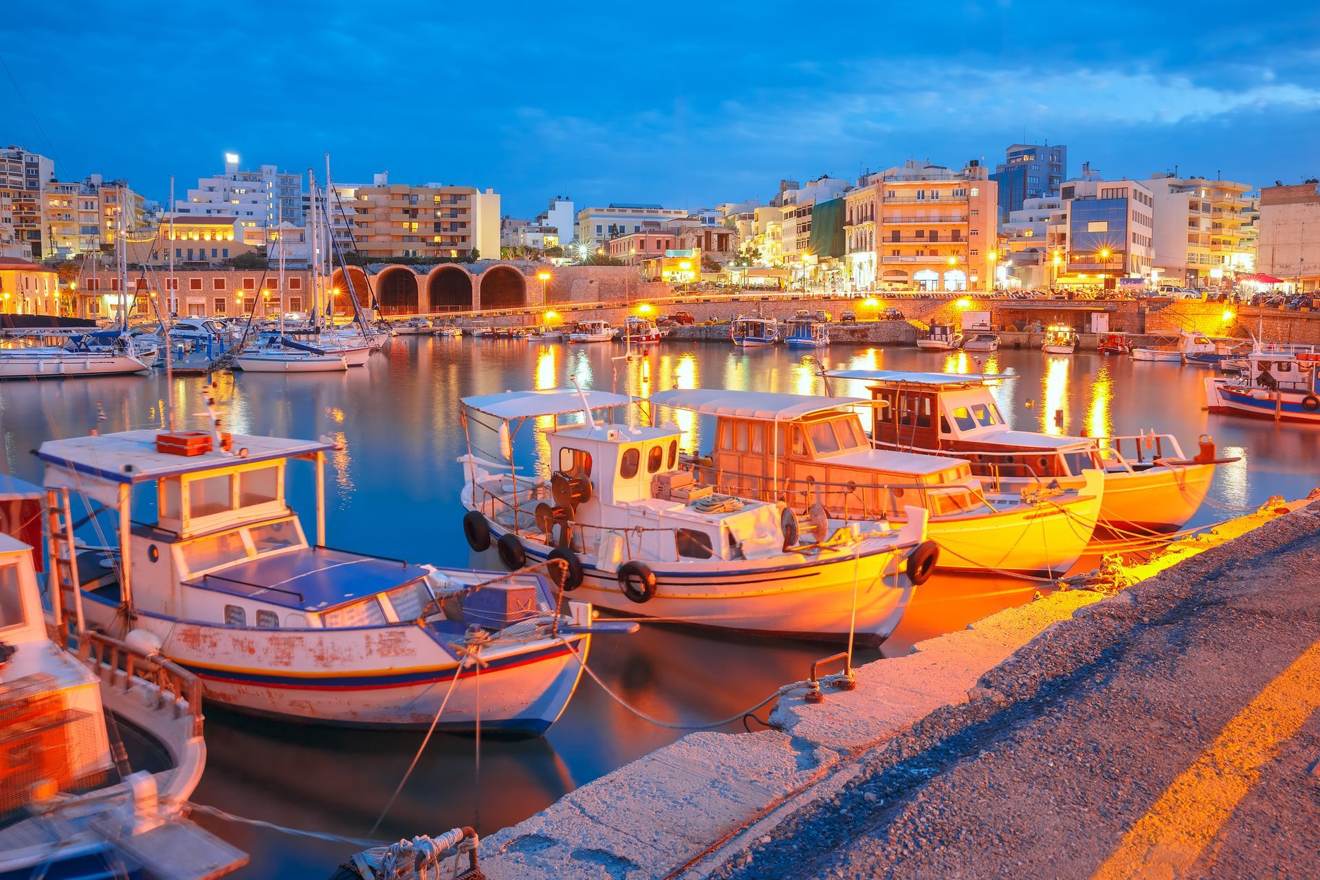 Boats docked in a harbor at dusk; buildings with lit windows in the background.