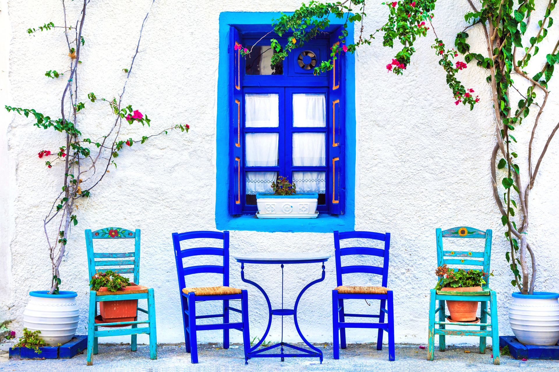 White building with a blue window and matching table and chairs, adorned with flowers in pots.