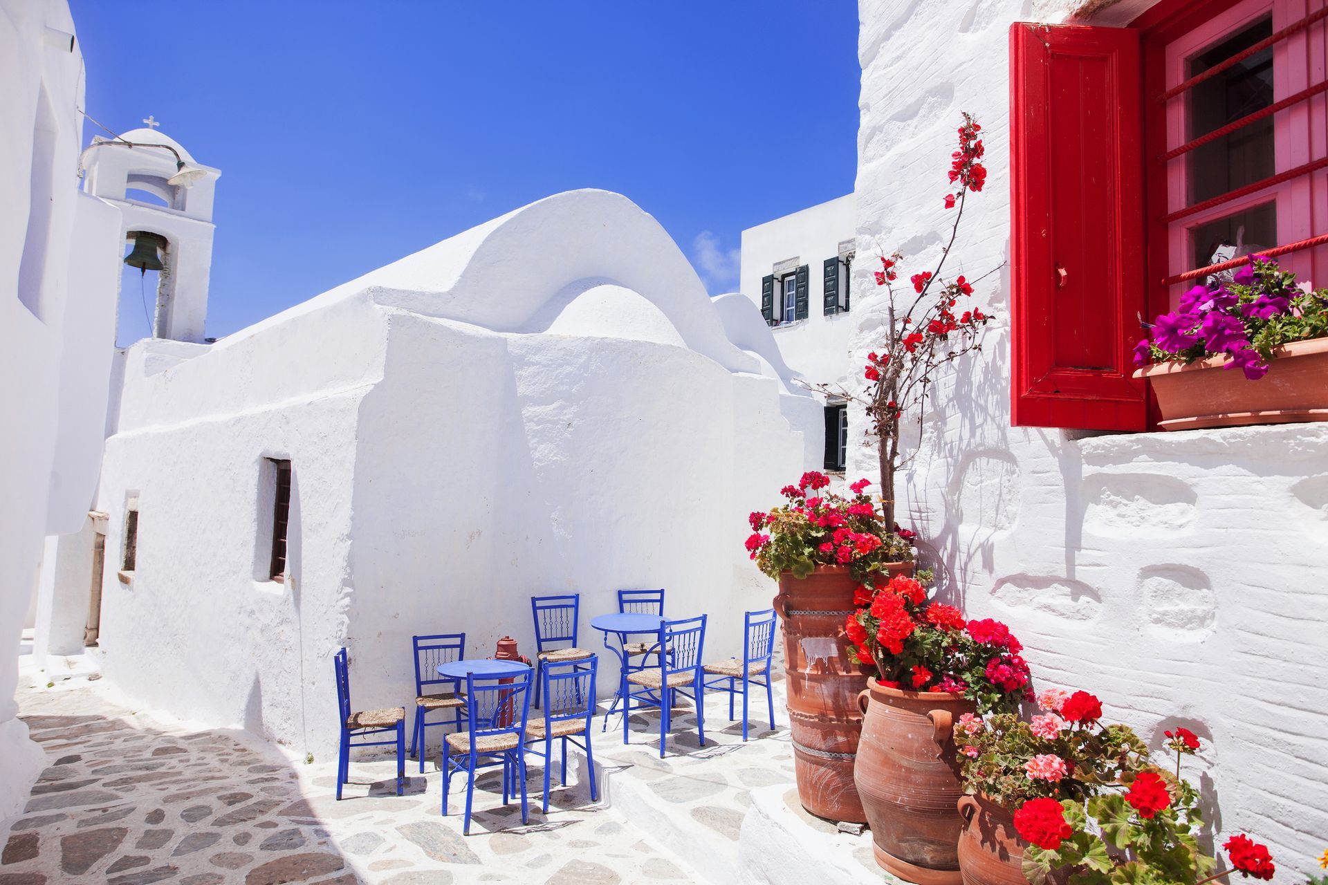 Whitewashed buildings with blue chairs and red flowers in a sunlit Greek alley.