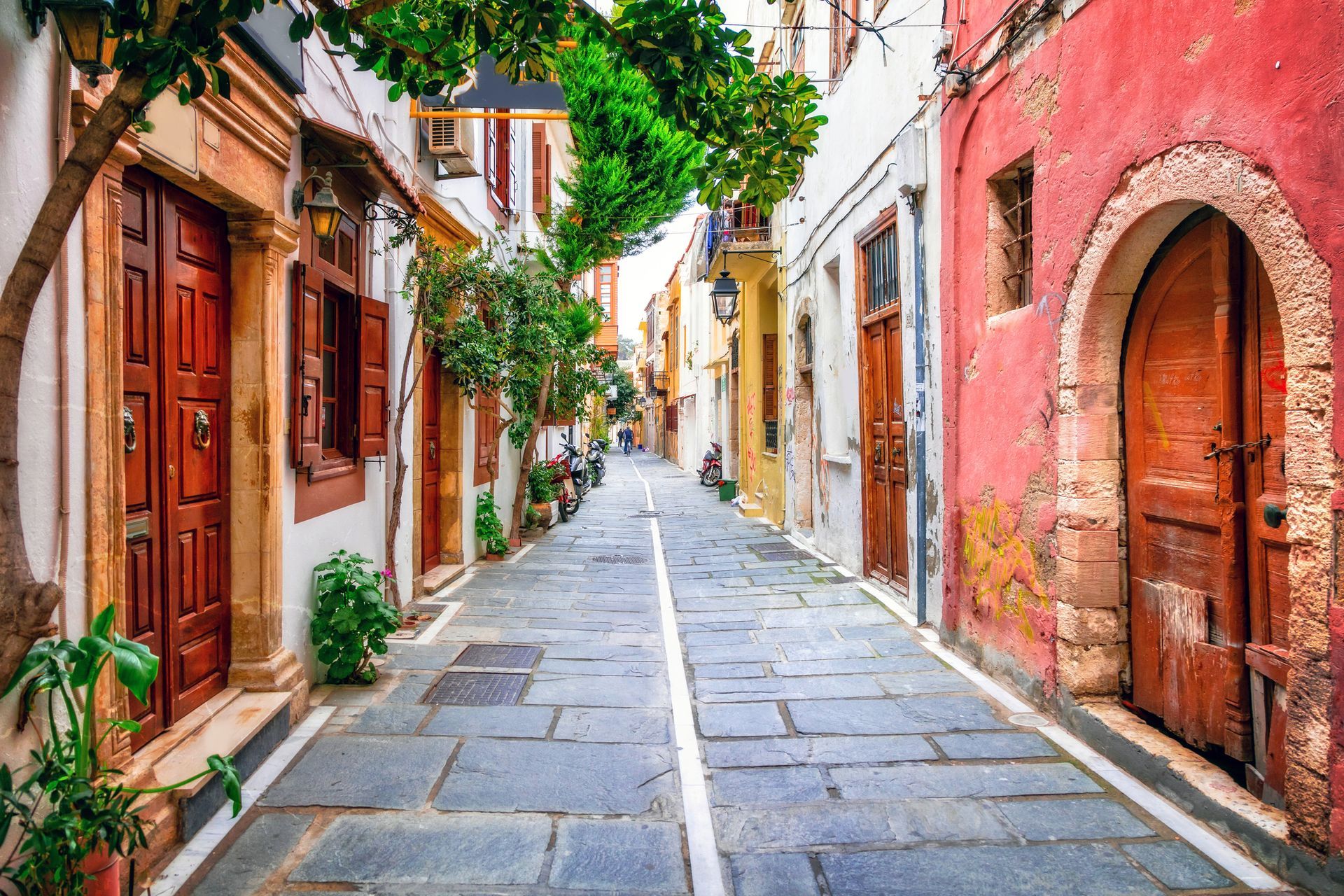 Narrow cobblestone street lined with colorful buildings in Greece. Arched wooden doors, trees, and sunlight.