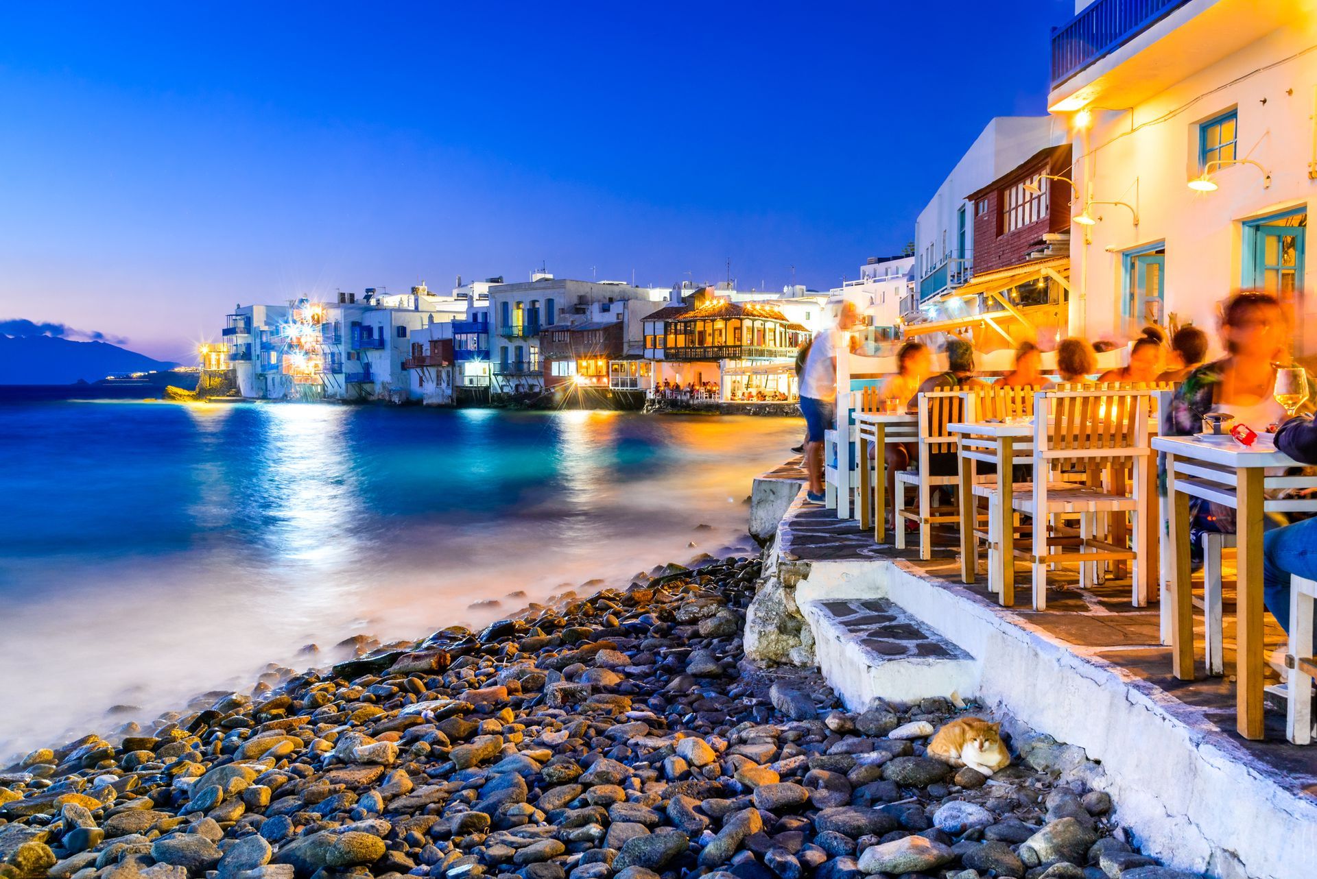 Coastal restaurant at dusk with people dining, illuminated buildings, and dark blue water.