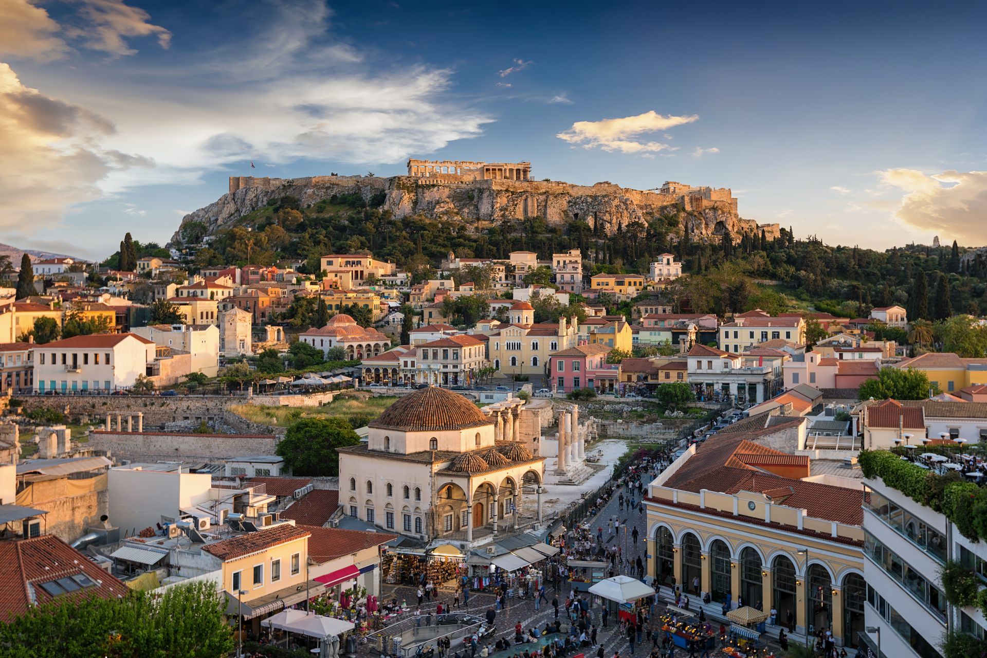 Athens, Greece: Cityscape with the Acropolis on a hilltop, buildings below, people in a plaza, blue sky.