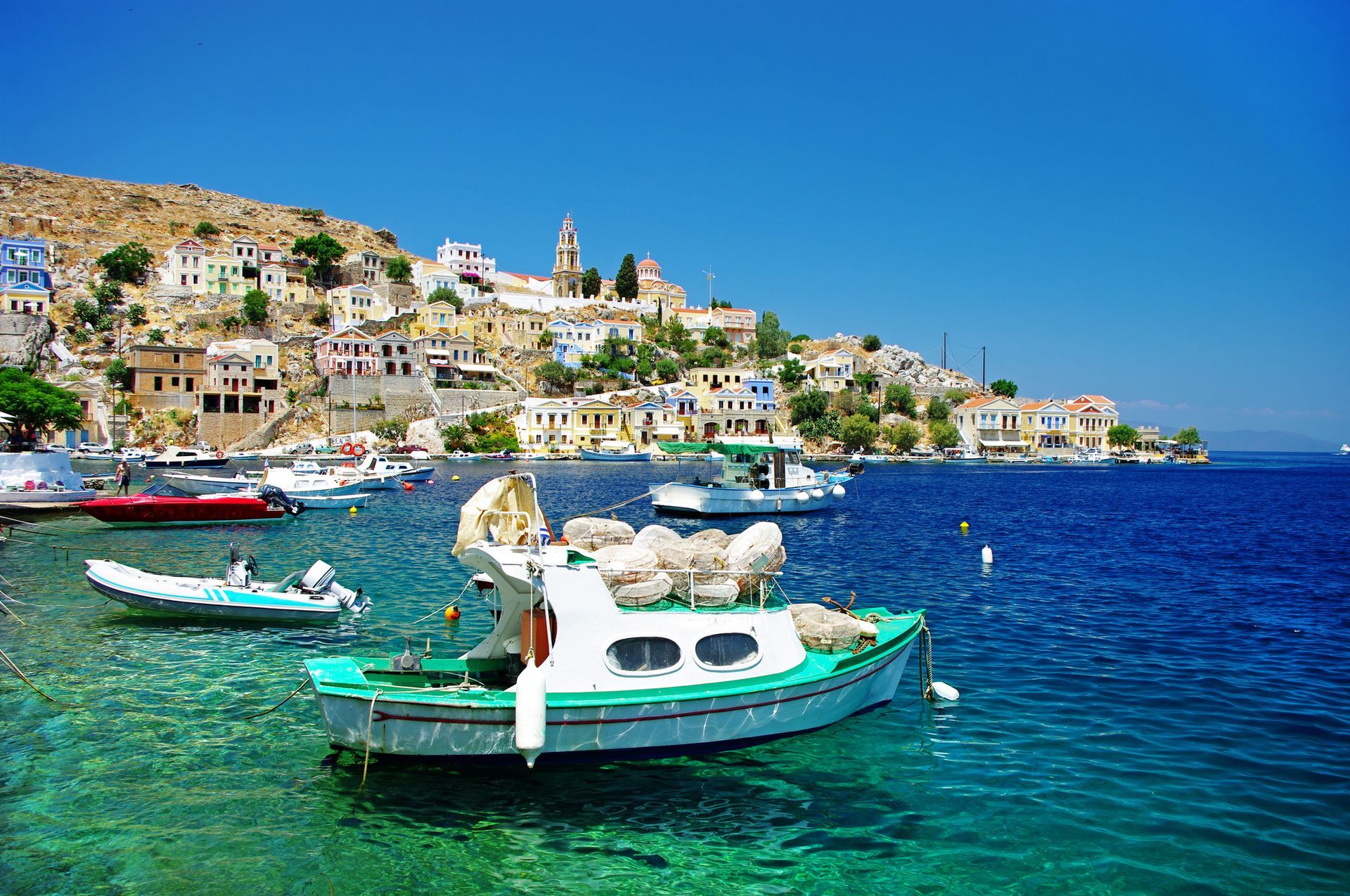 Boats in turquoise water, harbor town with colorful buildings on a hillside under a blue sky.