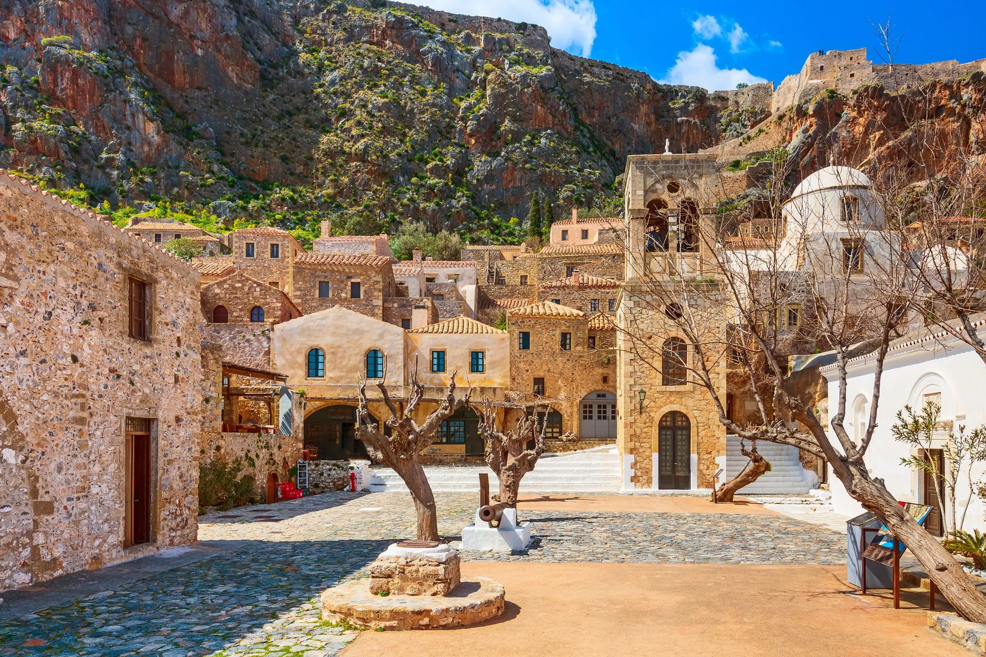 Medieval stone buildings in a sunlit square with a bell tower and mountainous backdrop.