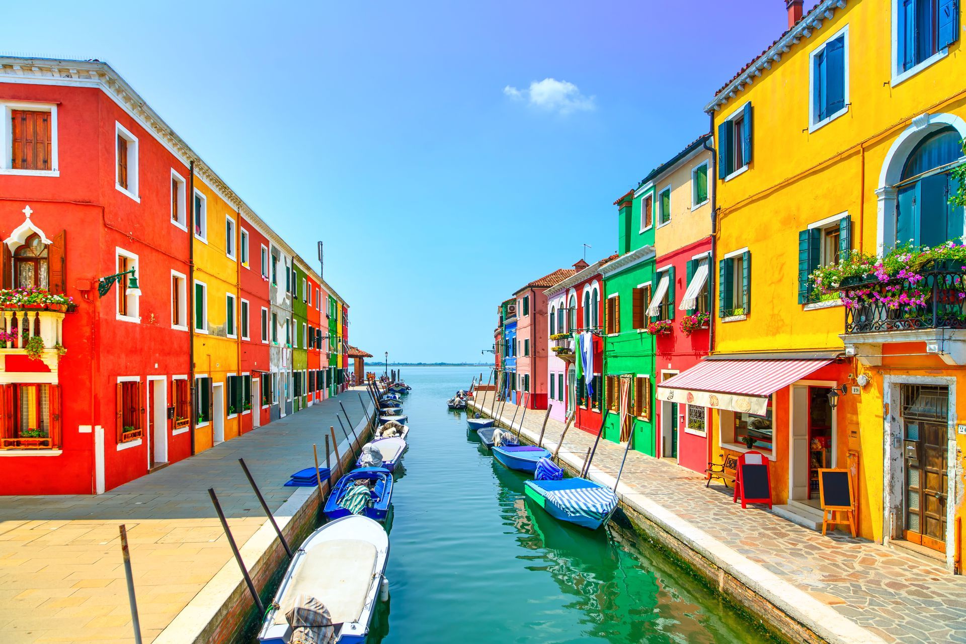 Colorful buildings line a canal with boats in Burano, Italy, under a clear blue sky.