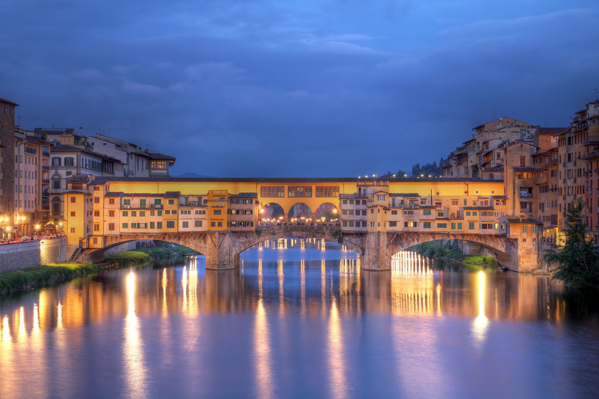 Ponte Vecchio bridge in Florence, Italy, illuminated at dusk, reflecting in the Arno River.