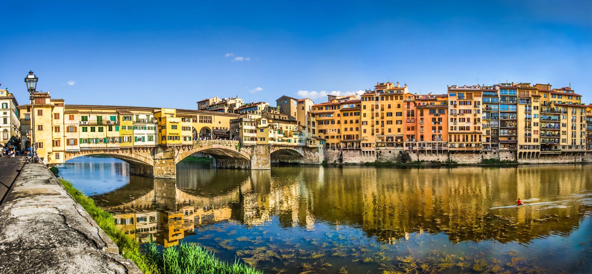 The Ponte Vecchio bridge in Florence, Italy, over a river, with buildings and reflections in the water under a blue sky.