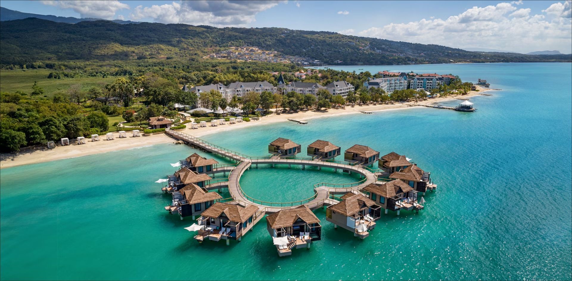 Overwater bungalows in turquoise water off the coast. Buildings on the shore in the background.