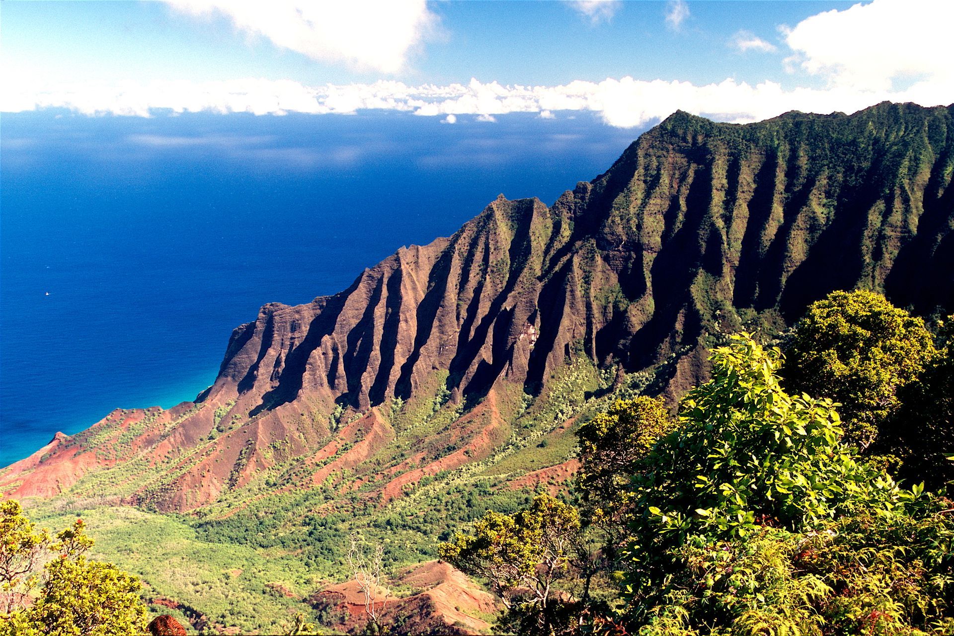 Rugged green and brown mountain range meets blue ocean, under cloudy sky.