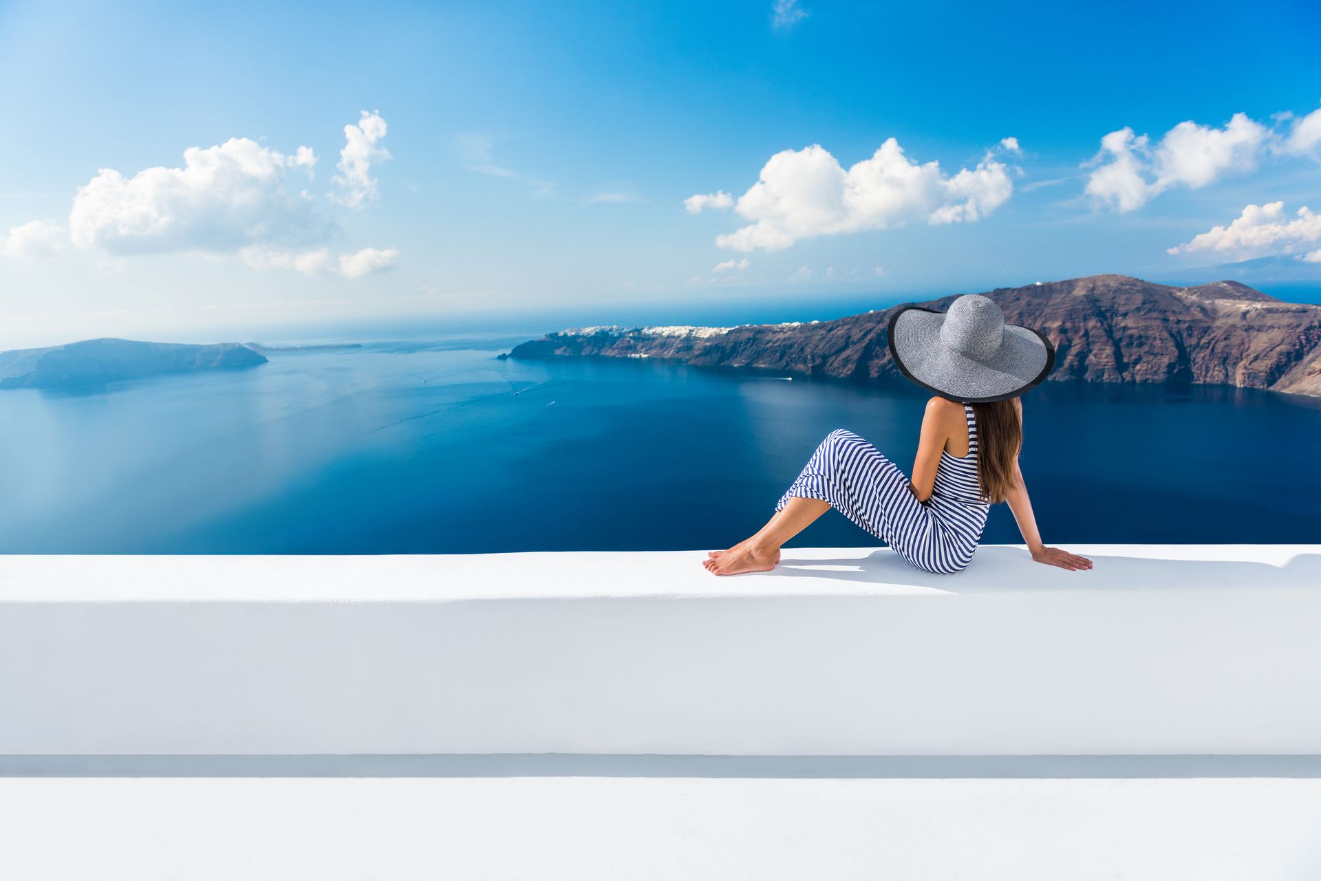 Woman in hat and striped outfit sitting on a white ledge, gazing at blue sea and islands.