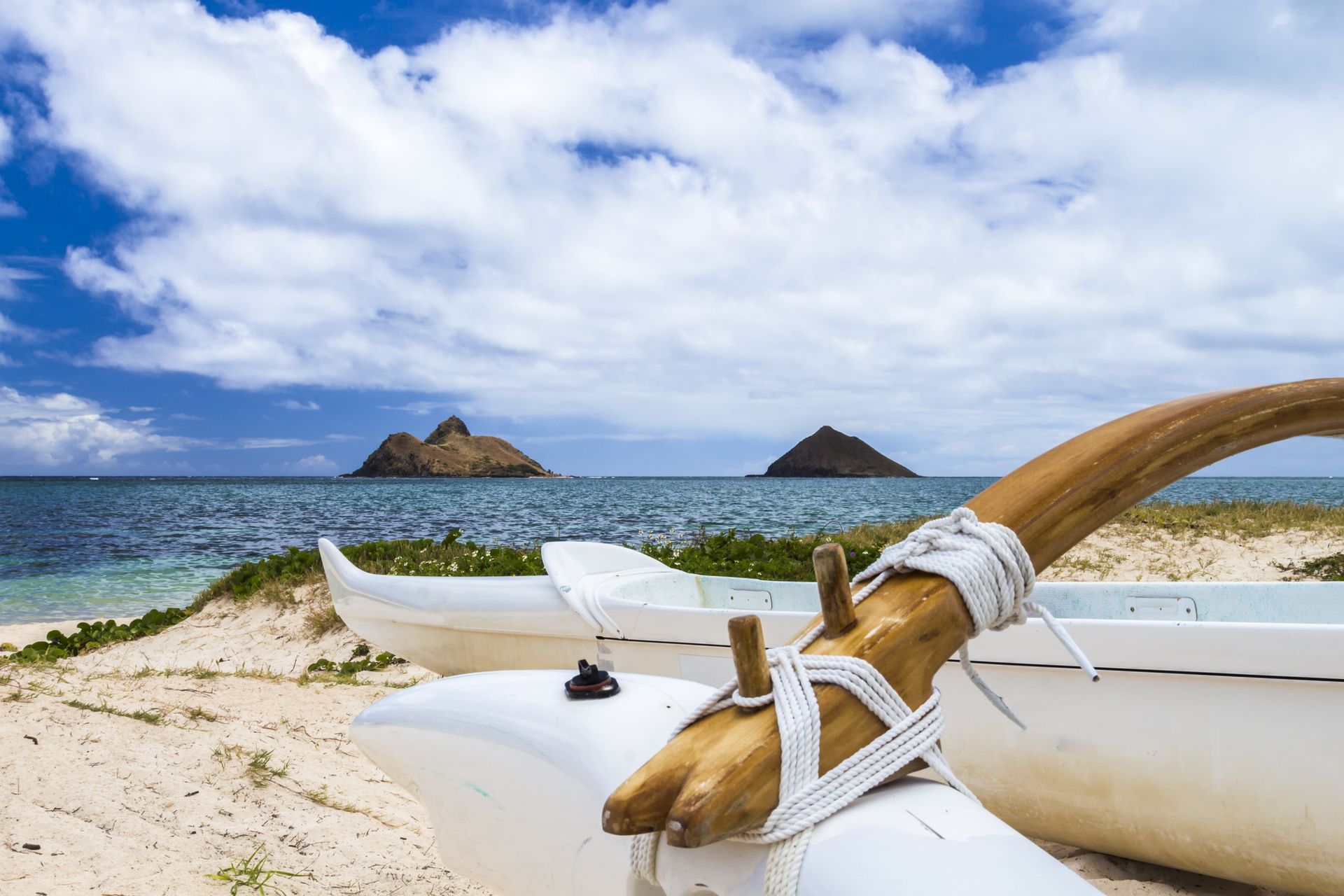 White outrigger canoe on sandy beach, two small islands in the distance, blue ocean and cloudy sky.