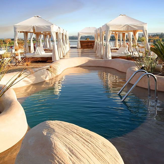 Pool with cabanas, sun loungers. Light-colored, tropical setting with blue water, sunny.