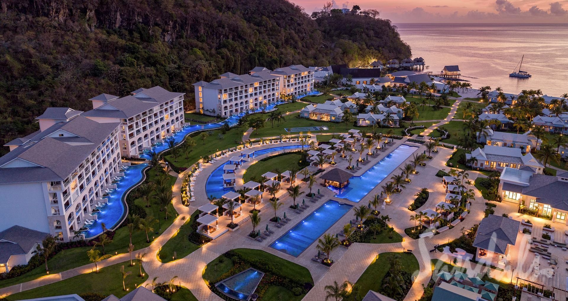 Aerial view of a resort with multiple white buildings, pools, and tropical landscaping, near a coast.