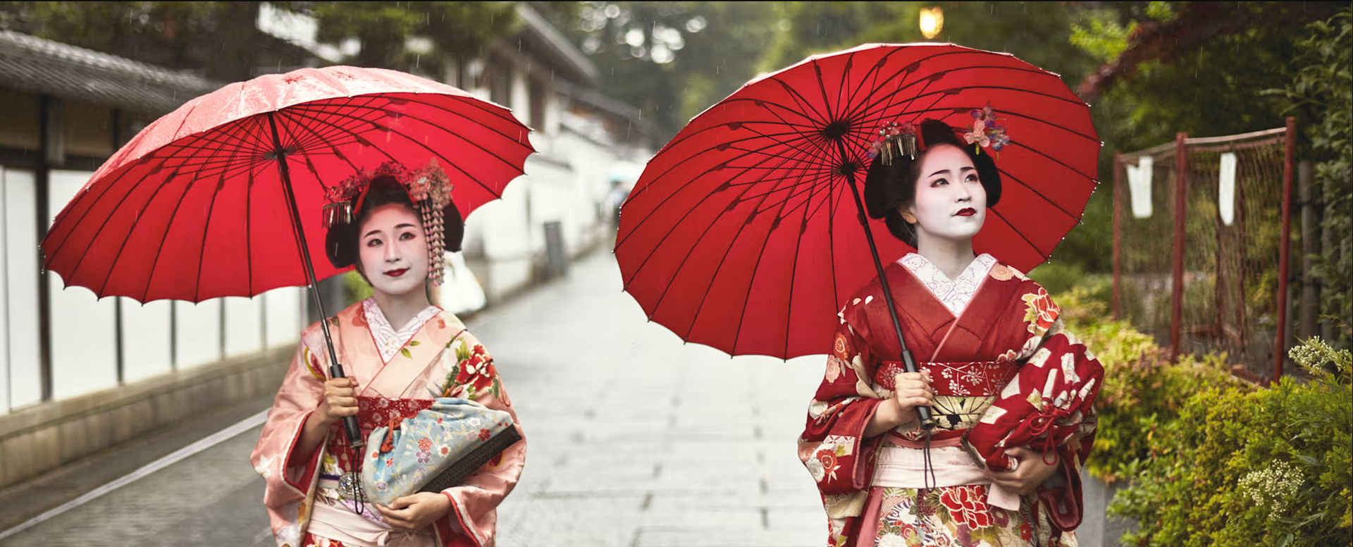 Two geishas with red umbrellas walking down a stone path in Japan.