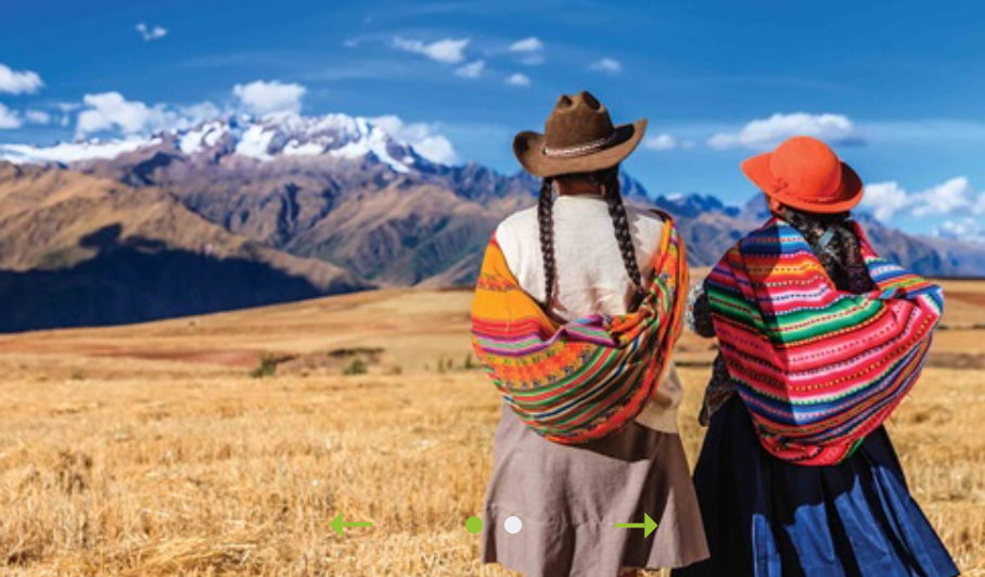 Two women in traditional clothing overlooking a mountain range, in a field.