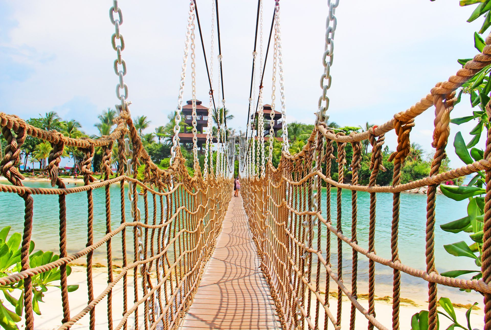 Suspension bridge over turquoise water, framed by greenery. Sandy beach and trees in the background.