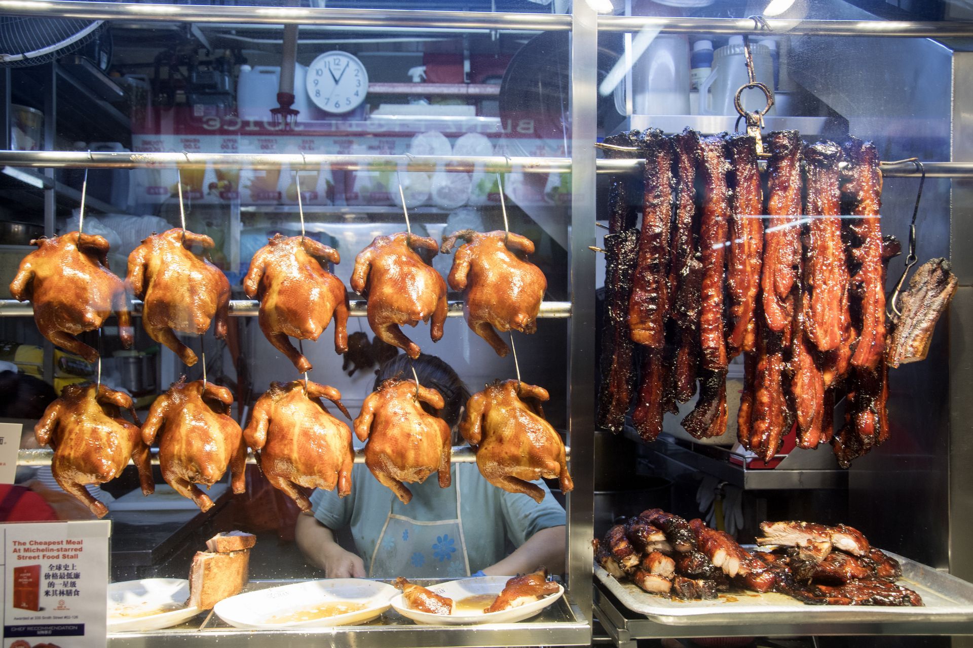 Roasted chickens and meats hanging in a restaurant window. A person is visible behind the counter.