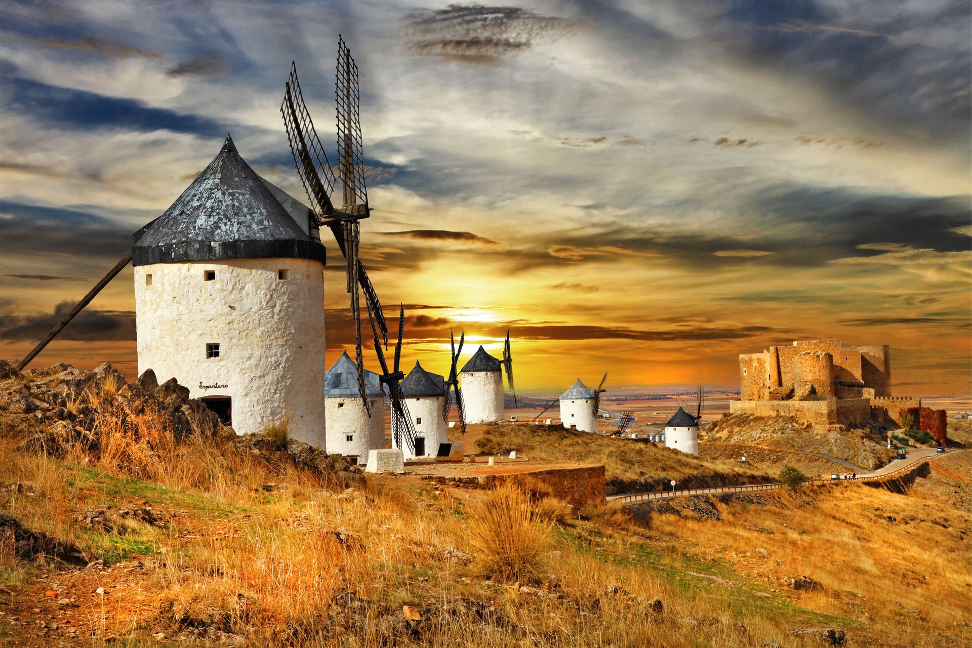 Windmills on a hill at sunset; white stone structures with dark blades; golden-hued sky.