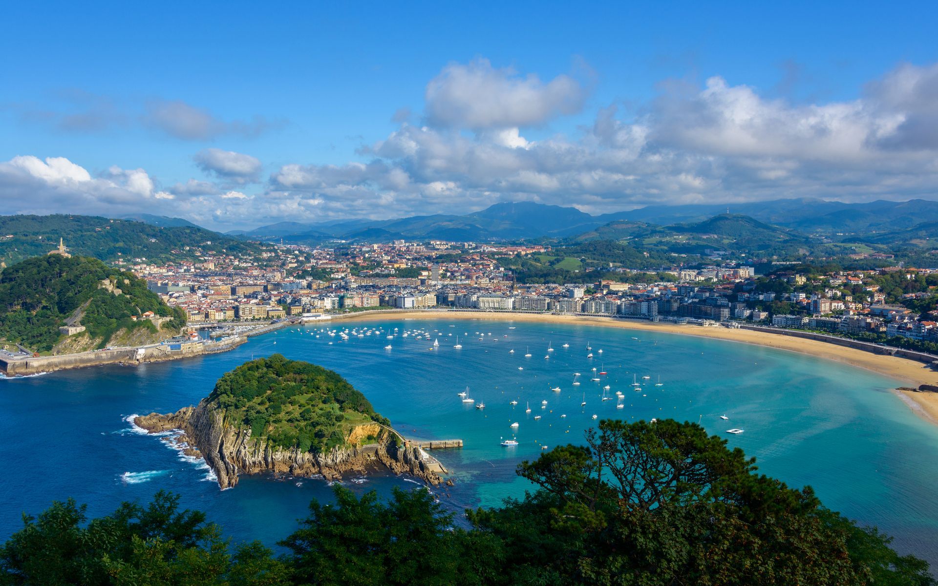 Panoramic view of San Sebastian, Spain, with a bay, boats, city buildings, and a small island under a blue sky.