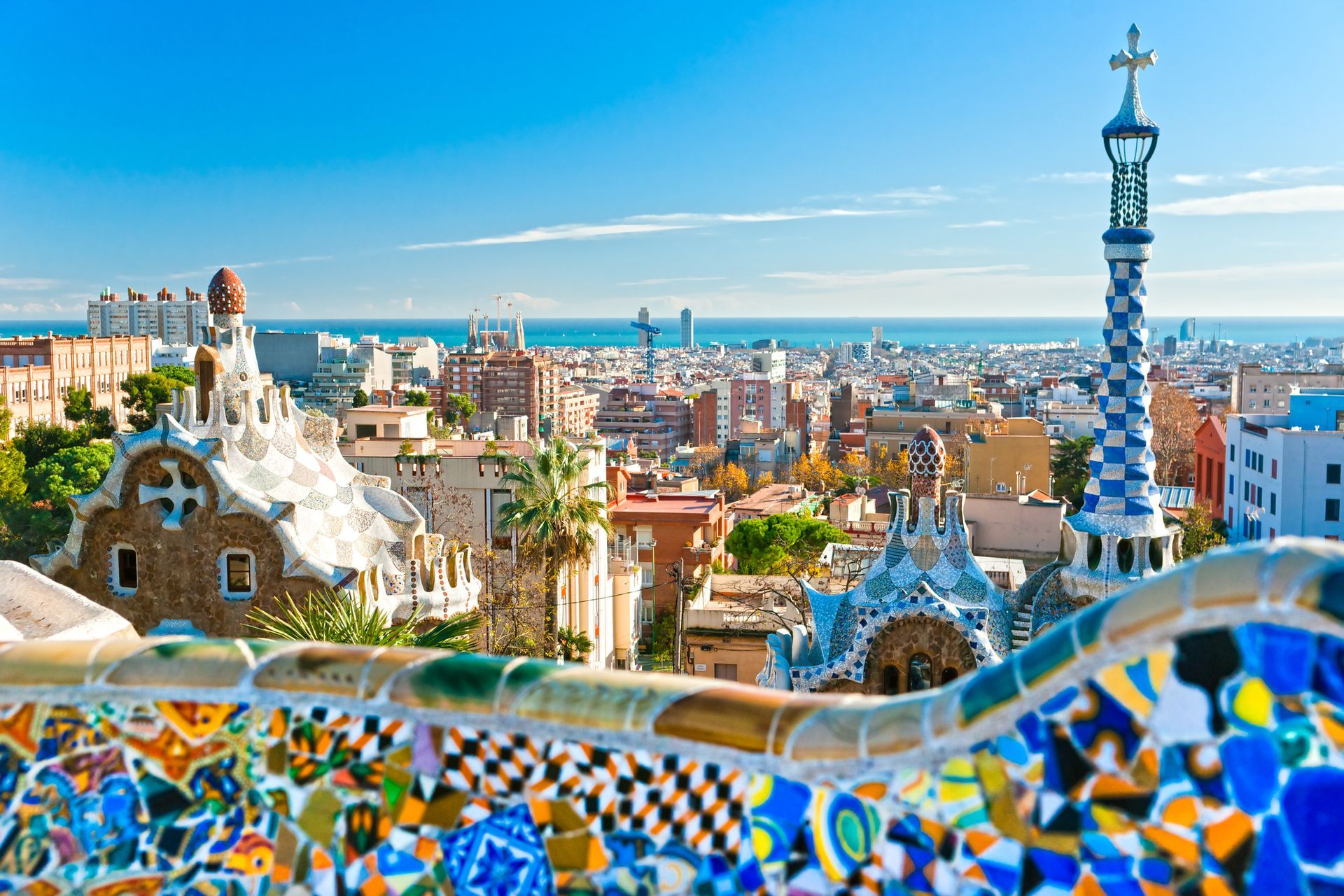 Barcelona cityscape from Park Güell, featuring colorful mosaic wall and tower, blue sky, and Mediterranean Sea.