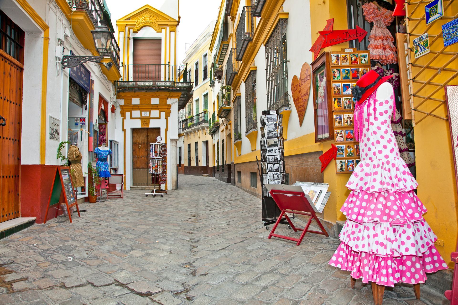 Cobblestone street lined with shops, displaying colorful dresses and souvenirs in Seville, Spain.