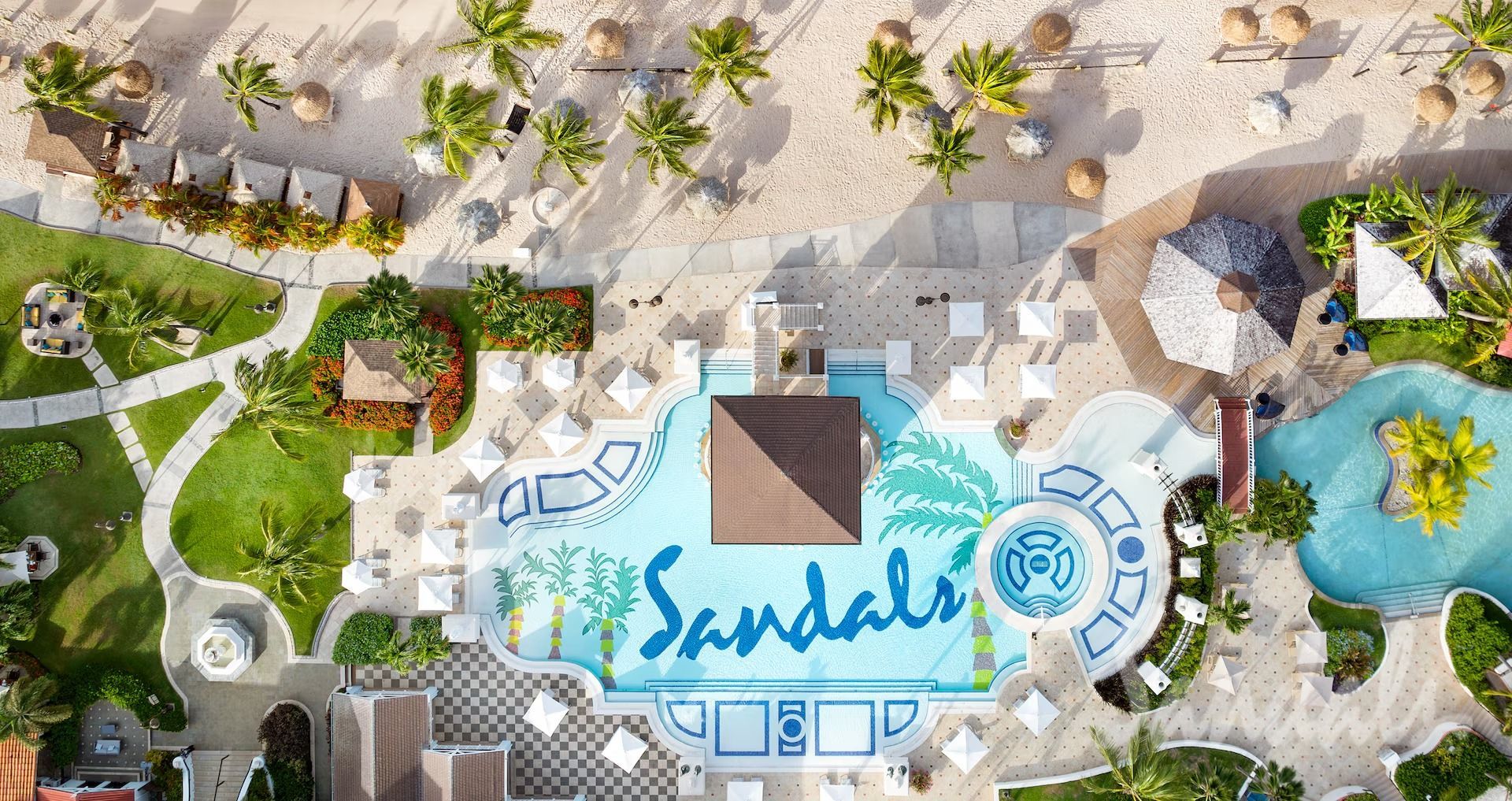Aerial view of a Sandals resort pool with the logo, beach, palm trees, and tropical landscaping.