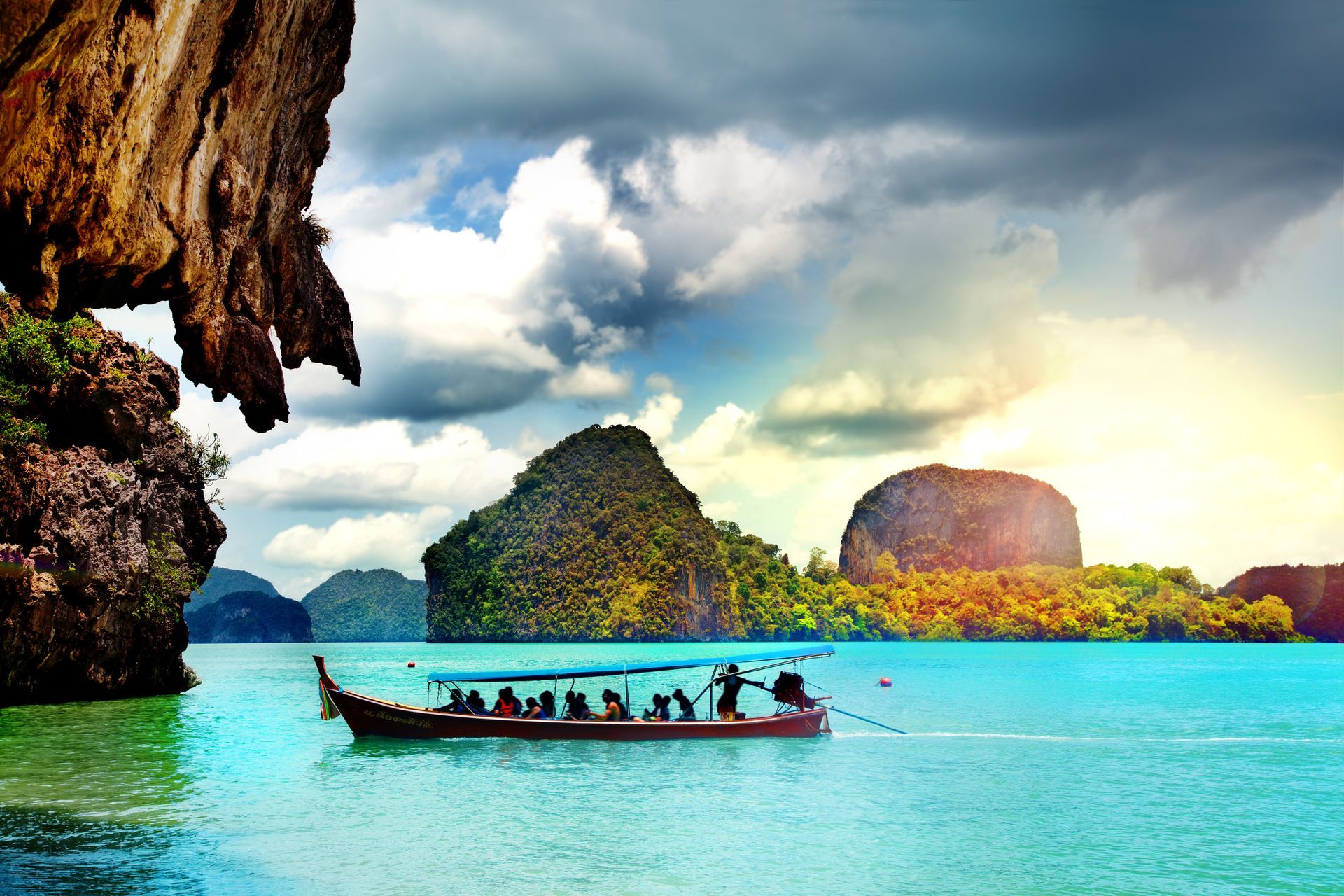 Longtail boat on turquoise water, with passengers, near limestone islands under cloudy sky.