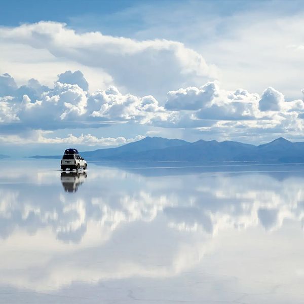 A white SUV driving on a reflective surface, mirroring the cloudy sky and distant mountains.