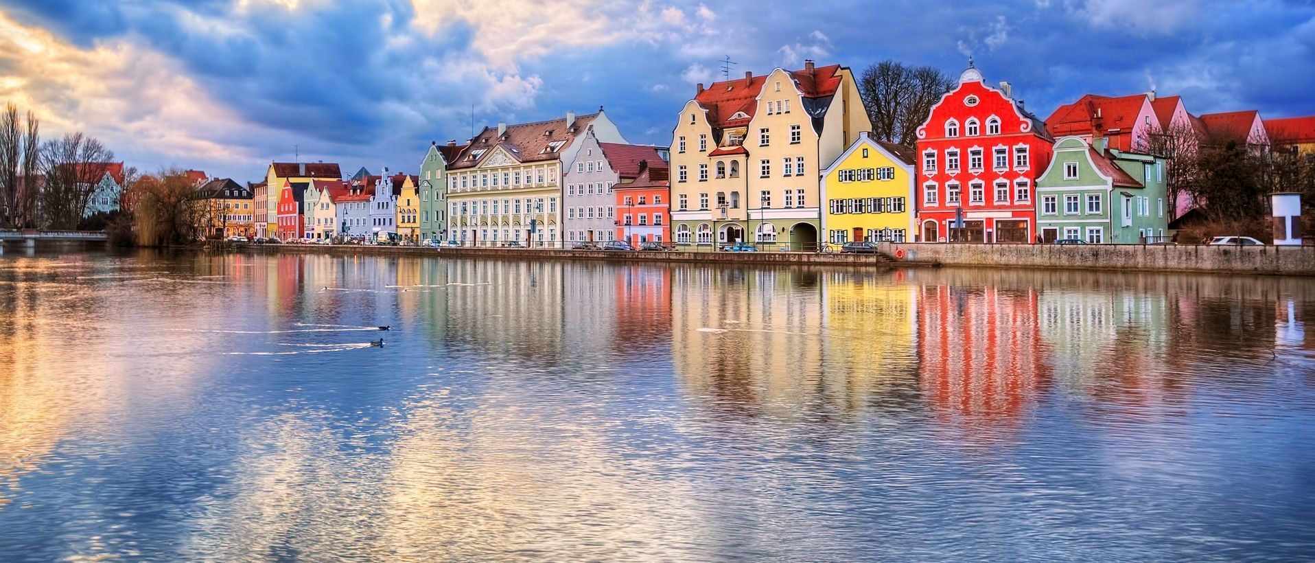 Colorful buildings reflect in the calm water, under a cloudy sky.