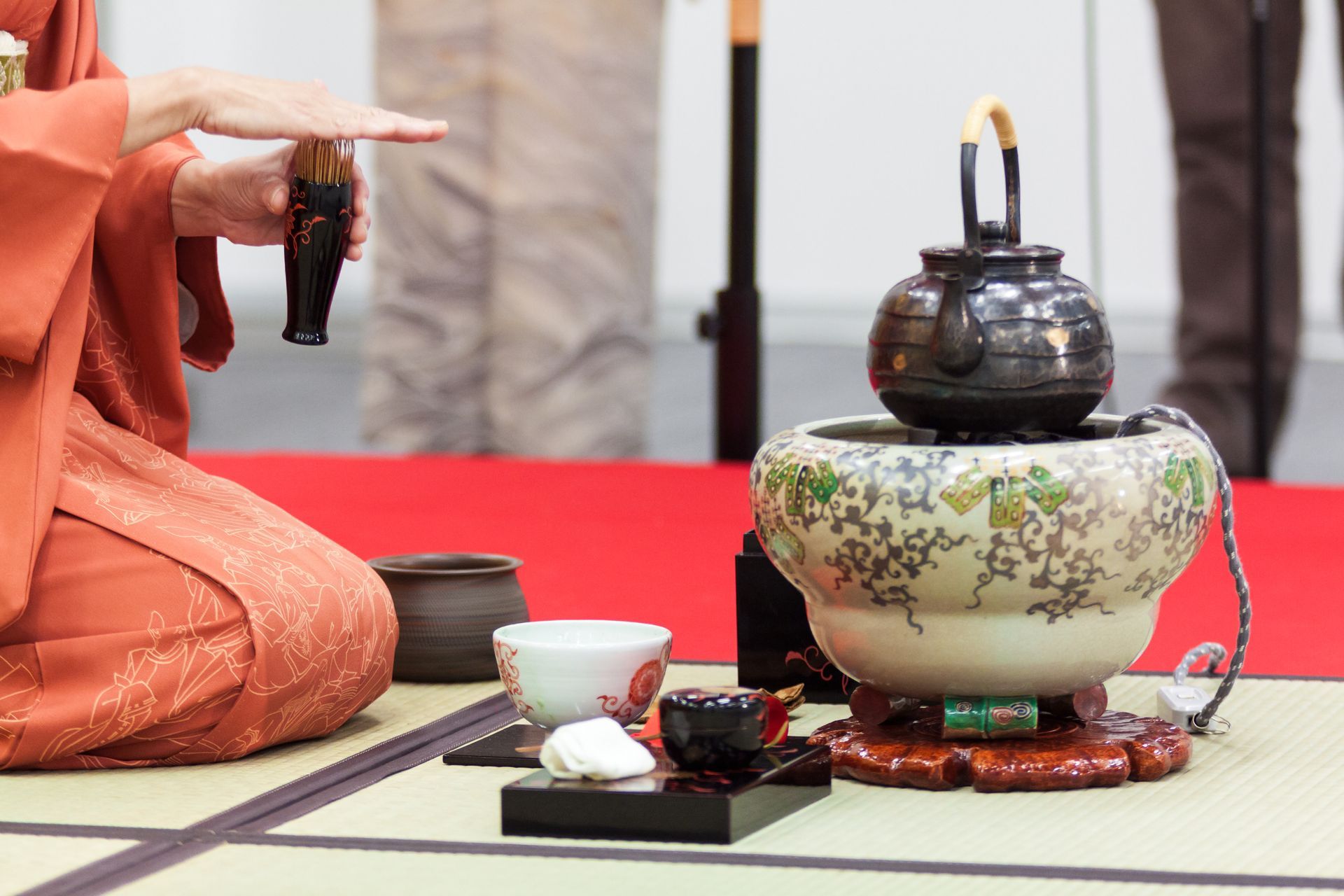 Person in kimono performing a Japanese tea ceremony, with utensils and teapot on a mat.