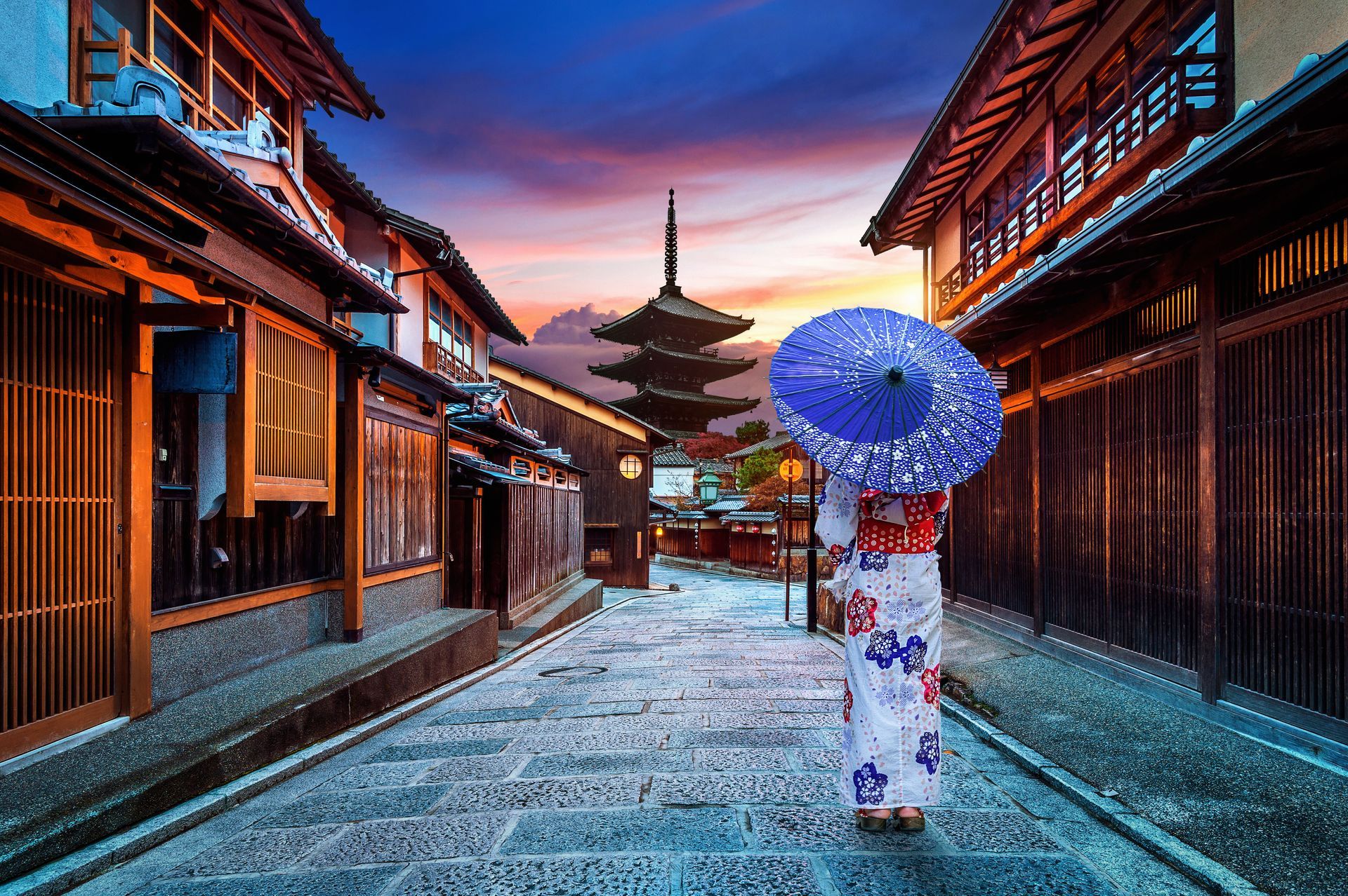 Woman in kimono holding umbrella on Kyoto street, traditional buildings, pagoda in background.