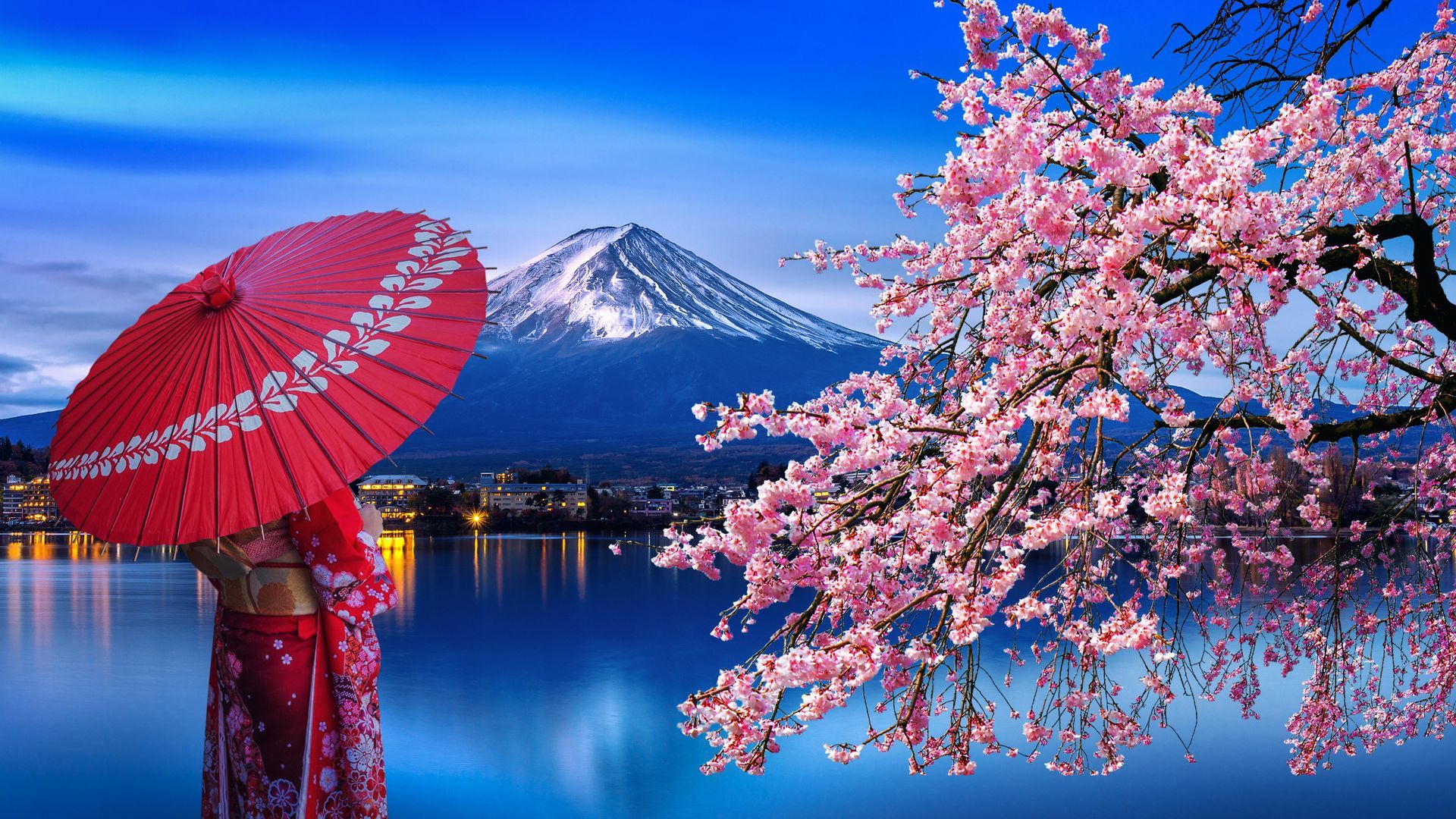 Woman in kimono holds red umbrella, Fuji mountain in background; cherry blossoms reflect in lake.
