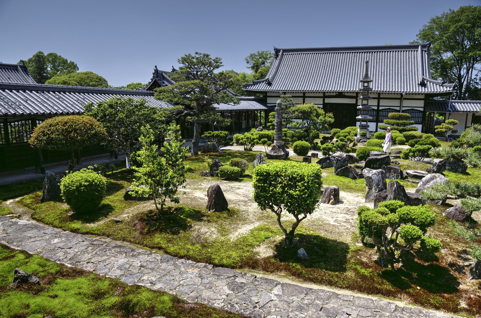 Japanese garden with trimmed shrubs, rocks, and a traditional building under a blue sky.